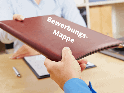 Eine Person übergibt eine braune Mappe mit der Aufschrift "Bewerbungs-Mappe" an eine andere Person auf dem Schreibtisch in einem Büro und weist damit auf das Unterstützungsangebot von Jobcoaching München hin.