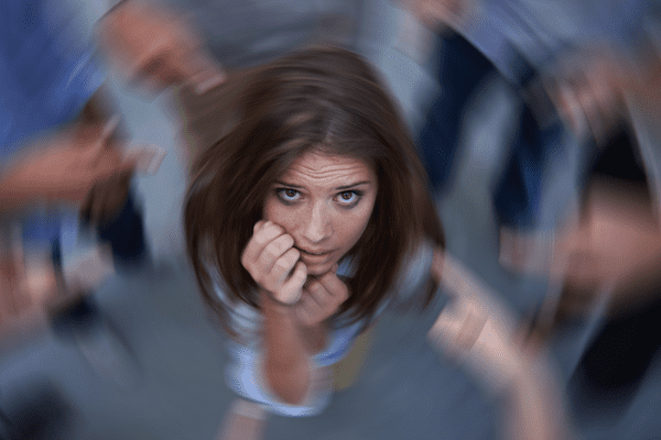 Eine junge Frau sieht ängstlich und verängstigt aus, umgeben von verschwommenen, zeigenden Händen. Das Bild ist mit einem radialen Unschärfeeffekt aufgenommen, der ihre Verzweiflung und ihr Gefühl, überwältigt oder beurteilt zu werden, unterstreicht.