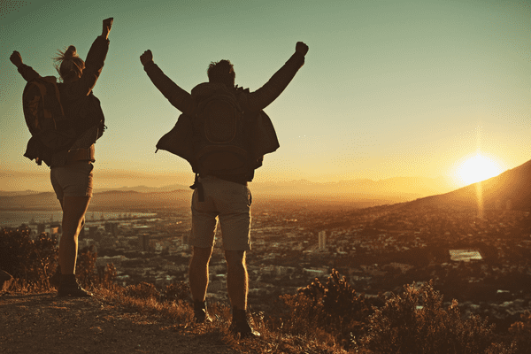 Zwei Wanderer mit Rucksäcken stehen bei Sonnenuntergang auf einer Bergkuppe und heben triumphierend die Arme, während sie die Stadt und die Landschaft unter sich betrachten, während die Sonne hinter den fernen Bergen untergeht.