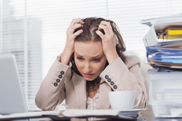 Eine Frau in Business-Kleidung sitzt an einem unübersichtlichen Schreibtisch und hält sich frustriert den Kopf. Umgeben von Papierkram und einem Laptop wirkt sie überfordert - vielleicht braucht sie Unterstützung wie das Jobcoaching München, um den Stress am Arbeitsplatz zu bewältigen.