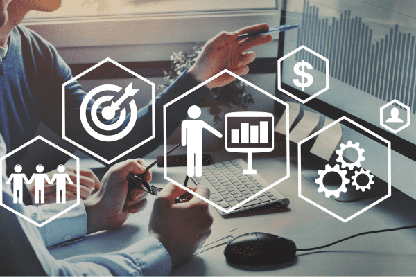 Zwei Personen arbeiten an einem Schreibtisch mit einem Computer, der Datendiagramme anzeigt. Overlay-Symbole stehen für Teamarbeit, Ziele, finanzielles Wachstum, Prozesseinstellungen und das Engagement des Publikums - ideal zur Veranschaulichung der Dienstleistungen von Jobcoaching München.
