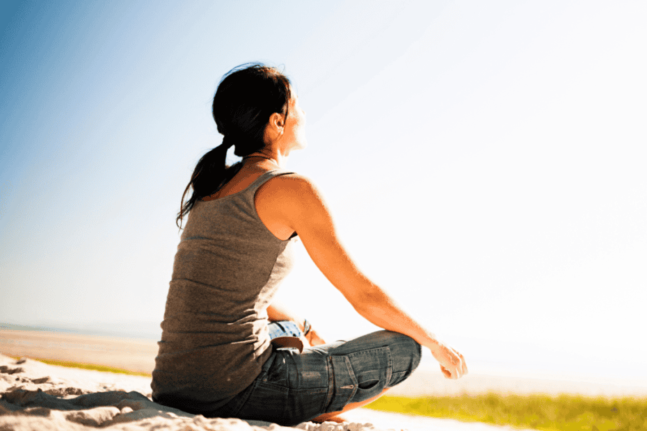 Eine Frau sitzt im Schneidersitz auf sandigem Boden und blickt mit ruhiger Miene in die Sonne. Der klare, helle Himmel sorgt für eine friedliche Stimmung. Der Text am unteren Rand lautet: "Die Kraft der Auszeit". Ideal für Jobcoaching München Momente der Selbstreflexion.