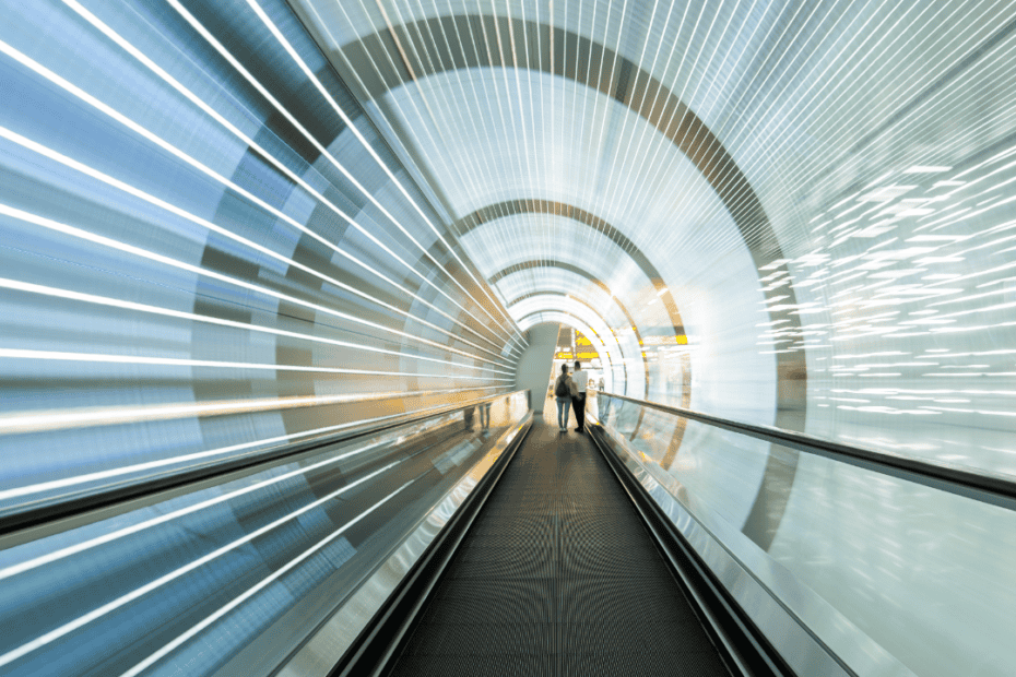 Ein futuristischer Tunnel mit Laufbändern, verschwommenen Lichtern und zwei Menschen, die in der Ferne gehen. Der große Text oben lautet "Elevator-Pitch" - ideal für Jobcoaching München-Profis, die auf der Suche nach Innovationen sind.
