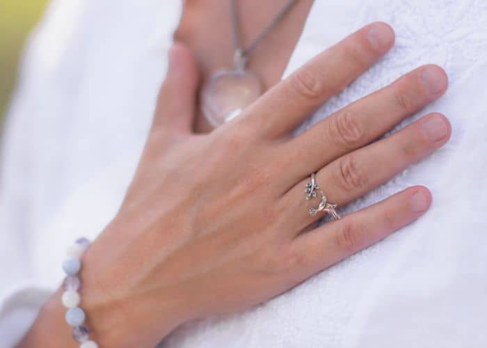 Eine Nahaufnahme einer Person, die ihre Hand über die Brust gelegt hat und ein Perlenarmband, silberne Ringe und eine Halskette mit Anhänger trägt, an der ein weißes, strukturiertes Kleidungsstück zu sehen ist - ein Zeichen für das Selbstvertrauen, das sie durch Jobcoaching München gewonnen hat.