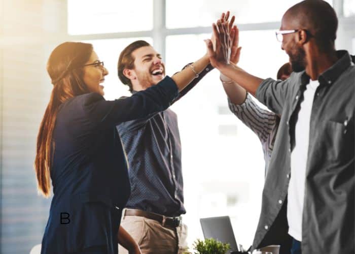 Vier Kolleginnen und Kollegen stehen in einem hellen Büro zusammen, lächeln und klatschen sich ab, um die Teamarbeit und den Erfolg von Jobcoaching München zu feiern.