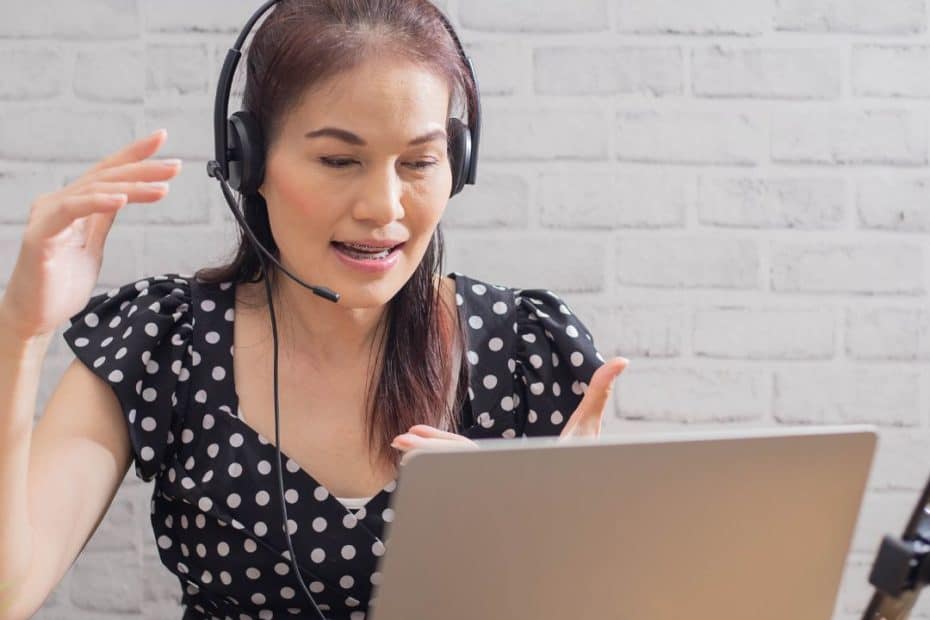 Eine Frau mit Headset sitzt an einem Schreibtisch mit Laptop, Notizbuch und Stift und gestikuliert während eines Videoanrufs. Der Text darüber lautet "Online-Vorstellungsgespräch" auf Deutsch - perfekt für Jobcoaching München.