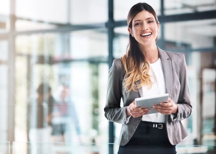 Eine Frau in Business-Kleidung steht in einem modernen B&uuml;ro, l&auml;chelt und h&auml;lt ein digitales Tablet. Der Hintergrund ist hell mit Glasw&auml;nden und unscharfen Figuren, die eine professionelle Atmosph&auml;re vermitteln.