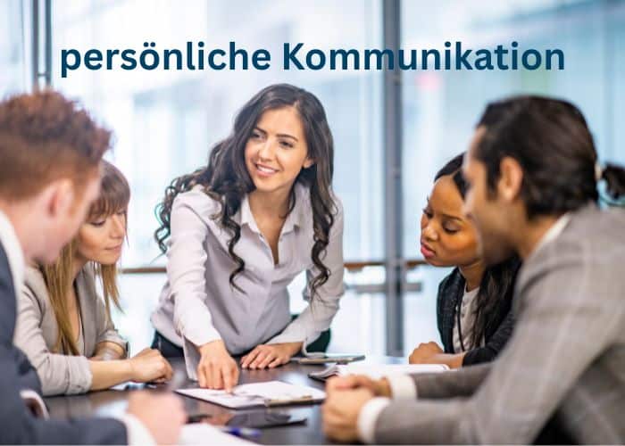 Eine Frau steht und deutet mit Gesten auf Dokumente auf einem Tisch, während vier Kollegen sitzen und aufmerksam zuhören. Über ihnen erscheint der Text persönliche Kommunikation.