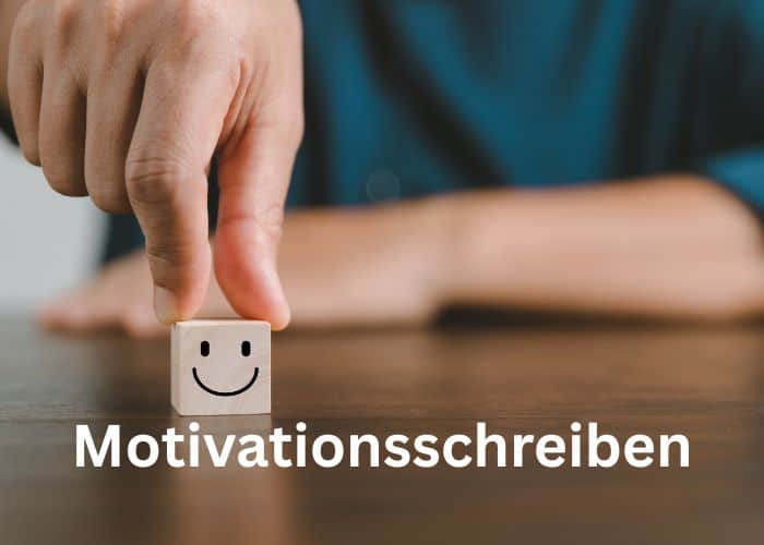 Eine Hand legt einen Holzblock mit einem Smiley-Gesicht auf einen Tisch. Das Wort Motivationsschreiben erscheint in fetter weißer Schrift am unteren Rand des Bildes.