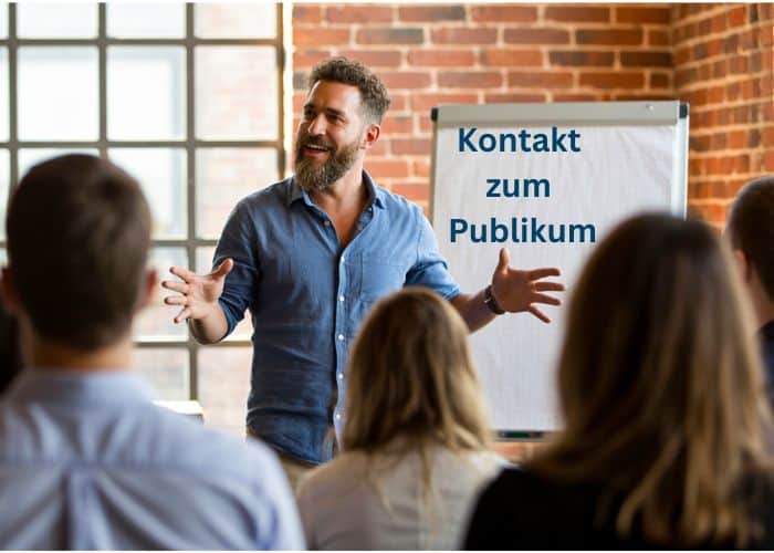 Ein Mann steht vor einer Gruppe, spricht und gestikuliert mit einem Lächeln. Hinter ihm steht ein Flipchart mit der Aufschrift "Kontakt zum Publikum".