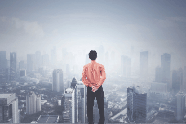 Ein Mann im roten Hemd steht mit den H&auml;nden auf dem R&uuml;cken und blickt von einem hohen Aussichtspunkt auf die neblige Stadtsilhouette - und denkt &uuml;ber neue Perspektiven nach, die er durch Jobcoaching M&uuml;nchen gefunden hat.